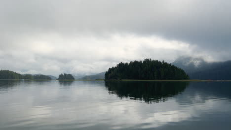 Wooded-Island-in-the-middle-of-a-lake-with-reflections,-moving-shot-from-a-boat,-moody-day,-Vancouver-Island
