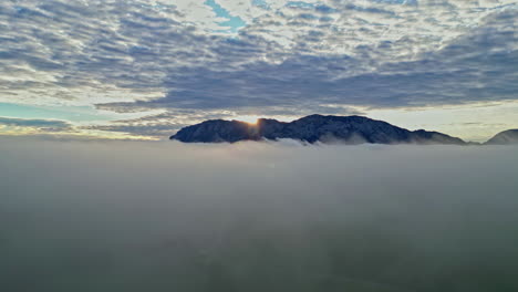 Aerial-view-of-sun-rising-behind-a-mountain,-piercing-through-a-blanket-of-clouds-at-dawn