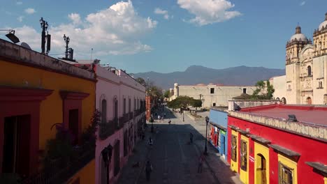 Luftflug-Zwischen-Bunten-Häusern-Mit-Gehenden-Menschen-In-Oaxaca-De-Juárez-Und-Der-Kathedrale-Im-Hintergrund