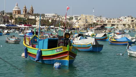 Colourful-Boats-Of-Marsaxlokk-With-The-Fishing-Village-In-The-Background