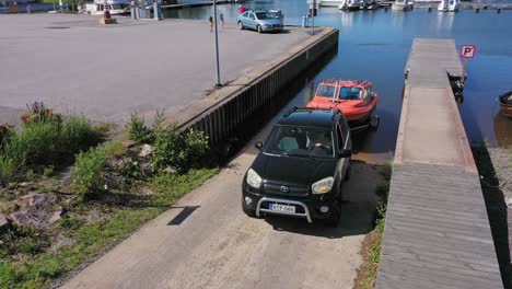 Aerial-view-of-car-and-trailer,-lowering-a-boat-at-a-marina,-lunching-it-into-the-water,-summer-day,-in-Finland---reverse,-drone-shot