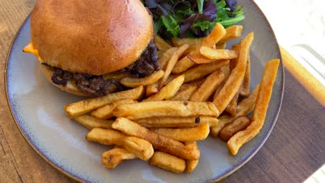 Tasty-cheeseburger-with-french-fries-and-salad-on-a-plate-by-the-sand,-beach-restaurant,-4K-shot