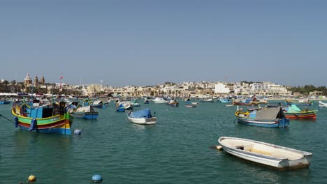 Coloridos-Barcos-Flotando-En-El-Puerto-De-Marsaxlokk-Con-Edificios-Al-Fondo