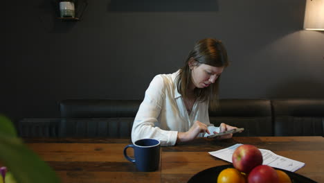 A-young-woman-texting-on-her-phone-in-a-kitchen-with-a-letter-and-paperwork-on-the-table