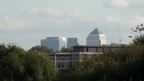 In-slow-motion-clouds-pass-Canary-Wharf-financial-district-in-the-distance-behind-a-residential-building,-as-trains-cross-between-two-groups-of-trees