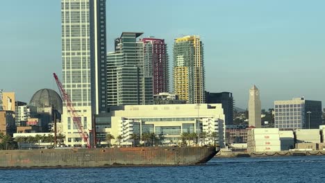 Rusty-tanker-cargo-ship-beneath-the-San-Diego-city,-California-skyline-in-Coronado-Bay