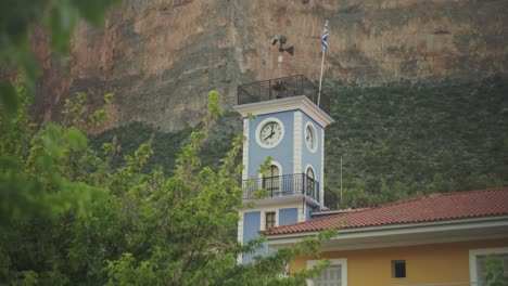 Vibrant-blue-clock-tower-with-Greek-flag-below-tall-cliffs-of-Leonidio-Greece