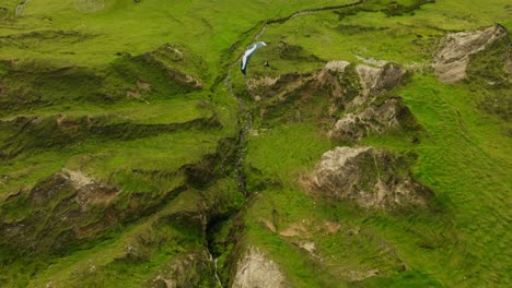 Paraglider-follows-mountain-stream-through-lush-mountainous-terrain,-aerial
