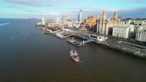 Mersey-Ferry-approaching-Liverpool-terminal-to-dock---tracked-by-drone-from-above-on-a-sunny-morning,-Liver-Buildings-backdrop