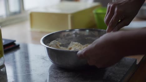 Gooey-dough-mixture-being-pressed-and-kneaded-by-spoon-in-steel-bowl,-filmed-as-close-up-slow-motion-shot-from-shadow-side