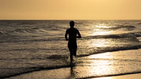 Young-man-running-along-the-beach-towards-the-camera-during-sunset