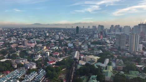 Aerial-flight-over-suburb-neighborhood-of-Manila-City-with-riding-train-and-skyscraper-buildings-in-background-at-sunset-time,-Philippines