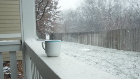 A-cup-of-hot-coffee-kept-on-a-balcony-railing-with-snow-falling-in-slow-motion-in-the-backyard-of-a-farmhouse-in-winter