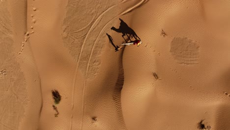 Die-Drohne-Fliegt-Von-Oben-Nach-Unten-Und-Folgt-Einem-Mann-Mit-Einem-Kamel-Und-Einer-Person-Oben-In-Der-Wüste,-Deren-Schatten-Auf-Dem-Boden-Liegt,-In-Tunesien,-Luftaufnahmen-4k