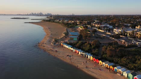 Drone-video-flying-over-Brighton's-famous-bathing-boxes,-with-Melbourne's-beautiful-City-skyline-in-the-far-horizon-at-sunset