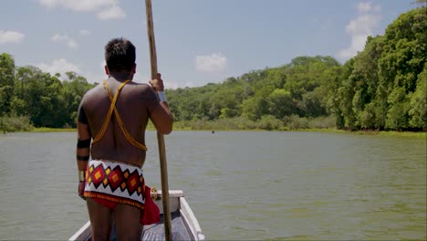 Tribal-man-standing-on-a-boat-it-a-river-with-Trible-costume-and-a-stick-in-hand-PANAMA,-CHAGRES-RIVER,-EMBERA-INDIAN-MAN-STANDING-AT-FRONT-OF-DUGOUT-CANOE