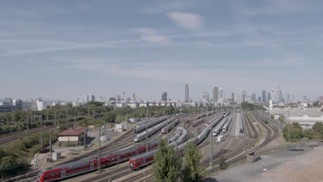 Eine-Weite-Luftaufnahme-Des-Frankfurter-Bahnhofsgeländes-Mit-Einer-Mischung-Aus-Zügen-Und-Der-Skyline-Der-Stadt-Im-Hintergrund-Unter-Blauem-Himmel