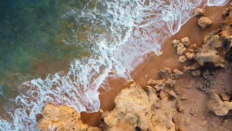 Aerial-top-down-rising-shot-of-foamy-waves-reaching-golden-and-rocky-beach-of-Portugal