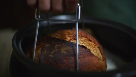 Hot-and-freshly-baked-bread-loaf-lifted-out-of-baking-pot-with-metal-tool,-filmed-as-closeup-slow-motion-shot