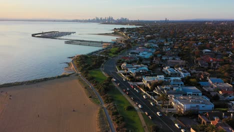 Drone-video-flying-over-Brighton's-beach-and-road-along-the-coast,-with-Melbourne's-beautiful-City-skyline-in-the-far-horizon-at-sunset