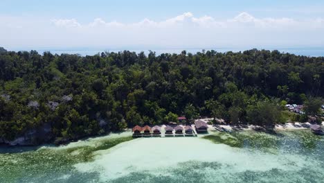 Vista-Aérea-En-Ascenso-De-La-Isla-Kri-Con-Cabañas-Frente-A-La-Playa,-Una-Densa-Selva-Tropical-Y-Un-Barco-De-Buceo-Turístico-Amarrado-En-Raja-Ampat,-Papua-Occidental,-Indonesia