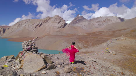 Tilicho-Lake-Manang-Nepal-landscape-reveals-woman-enjoying-in-pink-dress,-mountain,-cold-weather,-World's-Highest-Altitude-Lake-and-mountain-circuit-drone-shot-4K