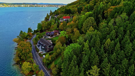 Aerial-road-and-waterfront-lakeside-houses-near-clear-freshwater-lake,-dense-green-forest
