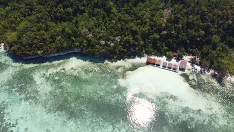 Steigende-Luftaufnahme-über-Strandhütten-Am-Wasser-Mit-Blick-Auf-Kristallklares-Meerwasser-Und-Korallenriff-Auf-Einer-Mit-Regenwald-Bedeckten-Insel-In-Raja-Ampat,-West-Papua,-Indonesien