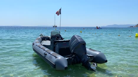 Inflatable-motor-boat-with-French-flag-floating-on-transparent-turquoise-sea-water-on-a-sunny-day-in-Cavalière-Lavandou-South-of-France,-fun-beach-holiday-vacation,-4K-static-shot