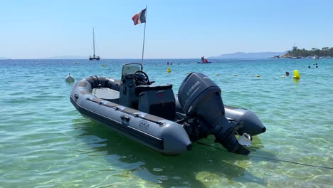 Inflatable-motor-boat-with-French-flag-floating-on-transparent-turquoise-sea-water-on-a-sunny-day-in-Cavalière-Lavandou-South-of-France,-fun-beach-holiday-vacation,-4K-shot