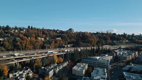 Drone-ascends-above-apartment-buildings-to-reveal-busy-highway-with-fall-foliage-lining-the-road