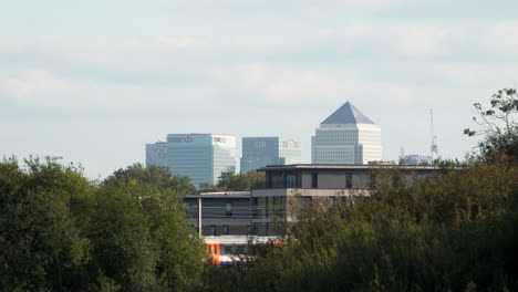 Canary-Wharf-financial-district-is-seen-in-the-distance-behind-a-residential-building,-as-a-train-crosses-between-two-groups-of-trees