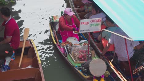 Mittlere-Aufnahme,-Frauen-In-Rosa-Schürze-Verkaufen-Gebratenes-Huhn-Und-Garnelen-An-Touristen-Auf-Dem-Schwimmenden-Markt-In-Thailand