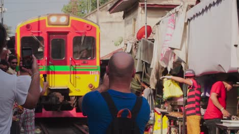 Medium-Shot,-Man-in-blue-shirt-Takes-photo-of-the-approaching-Train-in-Maeklong-Samut-Rail-Road-Market-Thailand