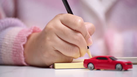Nahaufnahme-Einer-Hand,-Die-Einen-Bleistift-Hält-Und-Etwas-Auf-Gelbes-Papier-Schreibt,-Hinter-Einem-Roten-Spielzeugauto,-Das-Die-Finanzierung-Eines-Autokredits-Symbolisiert