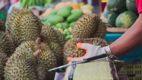 Focus-Shot,-Man-Slicing-a-jackfruit-in-half-in-Thailand-Market,-Assorted-fruits-blurred-in-the-background