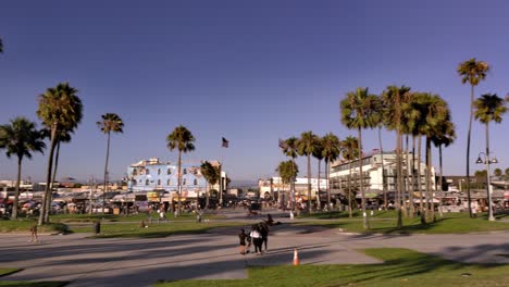 Venice-Beach-boardwalk-with-people-walking-around-and-riding-bikes-during-sunset-golden-hour-under-palm-trees-during-covid-19-pandemic---panning-shot
