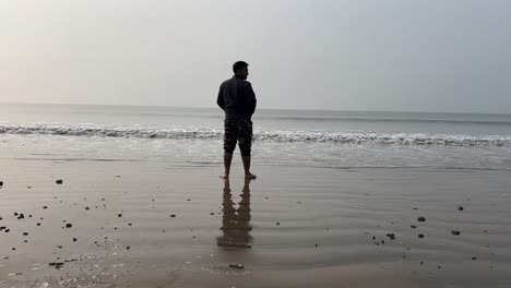 Rear-shot-of-a-man-standing-on-coastline-of-a-beach-with-small-waves-during-summer-in-Bengal,-India