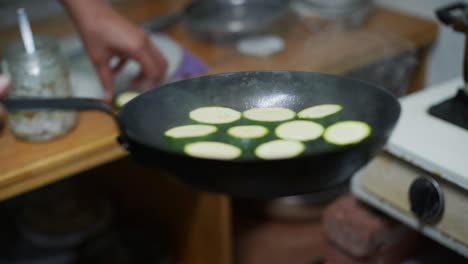 Cut-pieces-of-green-zucchini-placed-by-hand-in-black-fry-pan-with-hot-oil,-filmed-as-closeup-slow-motion-shot
