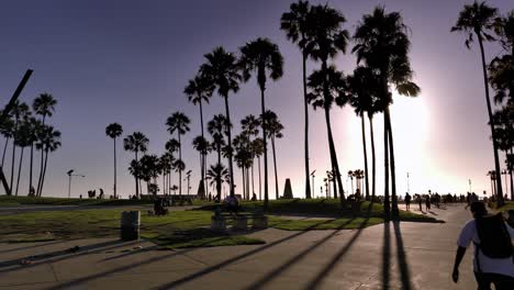 Venice-Beach-boardwalk-with-people-walking-around,-during-sunset-golden-hour,-under-palm-trees-and-shops-during-covid-19-pandemic---panning-shot