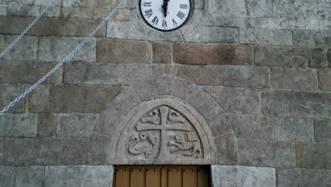 Heavy-wooden-doors-with-stone-coat-of-arms-and-clock-with-bell-tower-on-outside-of-church