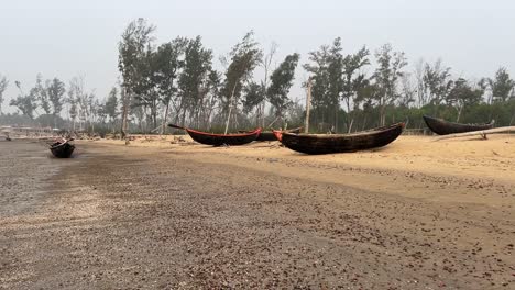 Wide-angle-shot-of-many-fishing-boats-kept-on-a-silent-empty-beach-in-daytime-with-some-trees-behind-them-in-Bengal,-India