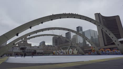 Verkleinern-Sie-Die-Ansicht-Von-Eisläufern,-Die-An-Einem-Bewölkten-Tag-über-Den-Nathan-Phillips-Square-Gleiten,-Mit-Dem-Rathaus-Von-Toronto-Im-Hintergrund