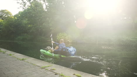Urban-kayaker-going-on-an-adventure-in-a-green-inflatable-kayak,-bright-sunlight-shines-through-trees,-slow-motion