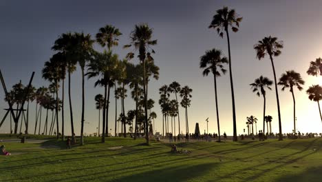 People-walking-around-Venice-Beach-boardwalk-during-sunset-golden-hour-under-palm-trees-during-covid-19-pandemic---panning-shot
