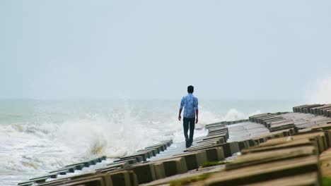 Man-appearing-to-be-depressed-and-lonely-walking-at-sea-shore-in-slow-motion-against-the-crushing-waves-on-a-concrete-breakwater-slope