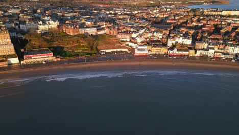 An-aerial-slow-motion-shot-of-busy-city-situated-beside-an-empty-beach-during-sunny-day-in-Scarborough-North-Yorkshire,-England