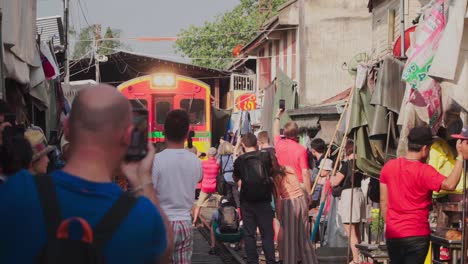 Establishing-Shot,-Tourist-Takes-Photo-and-Video-of-approaching-Train-in-Maeklong-Samut-Rail-Road-Market-Thailand