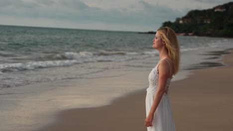 Blonde-girl-relaxing-on-the-beach-in-a-cute-white-dress,-beams-at-the-camera