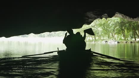 Silhouette-of-tourist-waving-traditional-Vietnamese-hat-boating-in-a-cave-at-Trang-An